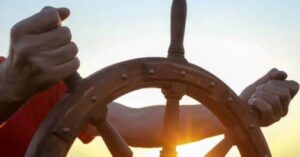 La foto muestra un timón de barco a la que se sujetan unas manos