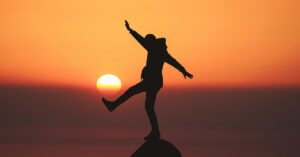 En la foto, una persona intenta equilibrarse sobre un promontorio, con el sol de fondo