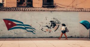 En la foto, una mujer cubana camina ante una pared con una bandera cubana y el logo borroso de la UJC