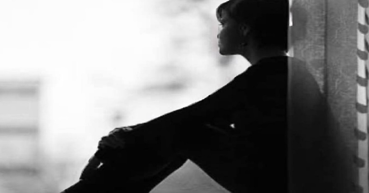 En la foto, una mujer aguada recostada a algo, hacia un exterior nebuloso