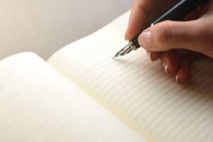 En la foto, una mano sostiene una pluma que escribe sobre un cuaderno