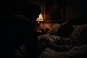En la foto, un hombre habla con un anciano sobre una cama en un cuarto en penumbras