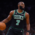 Fotografía deportiva en formato horizontal de un jugador de baloncesto de los Boston Celtics con uniforme negro y verde número 7. Sostiene el balón mientras sonríe ligeramente, con iluminación dramática que resalta su figura y un fondo oscuro y desenfocado que enfatiza su presencia en la cancha.