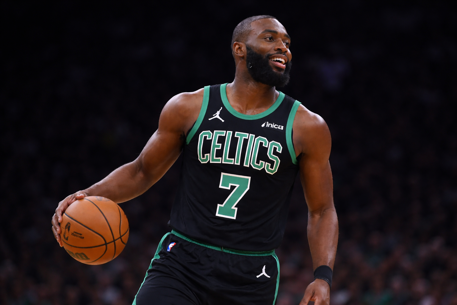 Fotografía deportiva en formato horizontal de un jugador de baloncesto de los Boston Celtics con uniforme negro y verde número 7. Sostiene el balón mientras sonríe ligeramente, con iluminación dramática que resalta su figura y un fondo oscuro y desenfocado que enfatiza su presencia en la cancha.