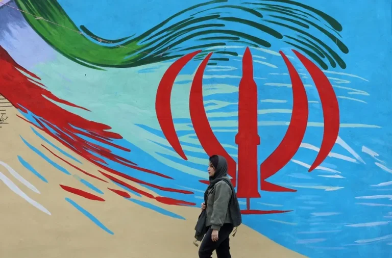 En la foto, una mujer caminando frente a un mural con la bandera iraní, en Teherán (Irán)