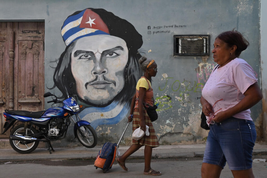 En la foto, dos personas pasan frente a un cartel con una foto del Che Guevara en La Habana