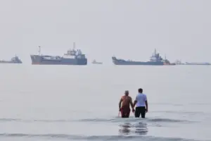 En la foto, Dos hombres vadean las aguas del estrecho de Ormuz con embarcaciones ancladas al fondo, frente a Bandar Abbas, Irán