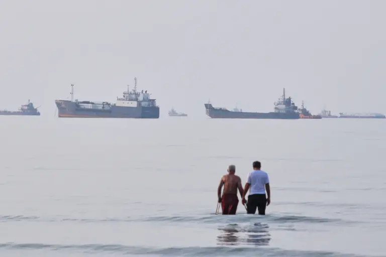 En la foto, Dos hombres vadean las aguas del estrecho de Ormuz con embarcaciones ancladas al fondo, frente a Bandar Abbas, Irán