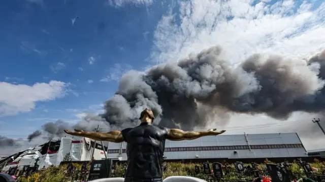 En la foto, una escultura funeraria en un cementerio mientras el humo se eleva sobre una instalación de Wildberries (al fondo) tras un ataque con drones ucranianos en el parque industrial de Koledino, cerca de Podolsk (región de Moscú, Rusia), el 16 de agosto de 2026
